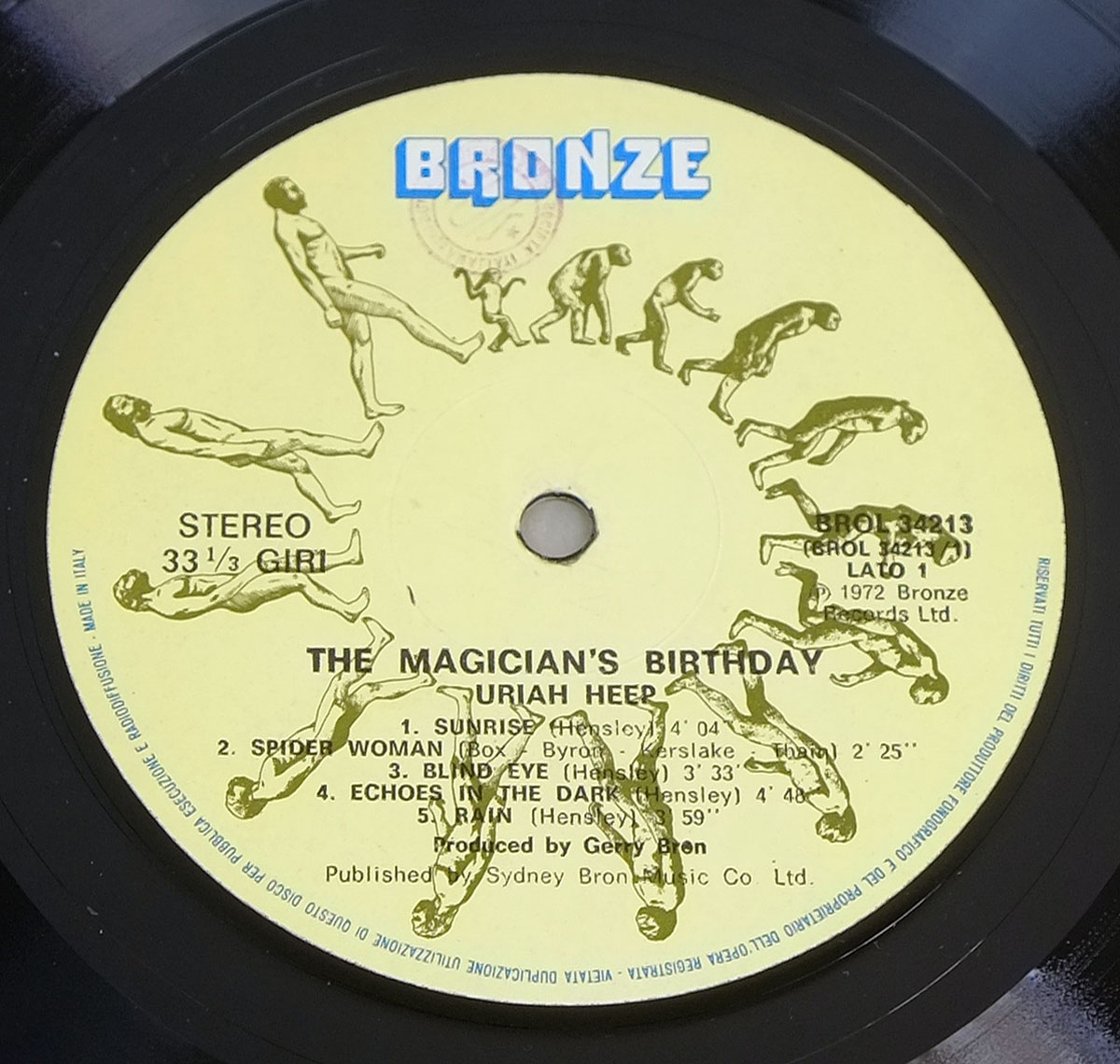 Bronze Records label of Uriah Heep's 'The Magician’s Birthday' vinyl LP. Features the iconic evolution sequence artwork depicting the progression from ape to modern man in a circular pattern. The label has a pale yellow background with a bold blue 'BRONZE' logo at the top. Text includes 'STEREO 33 ⅓ GIRI' on the left, catalog number 'BROL 34213' on the right, and production credits for Gerry Bron at the bottom. 'Made in Italy' is printed along the outer rim.
